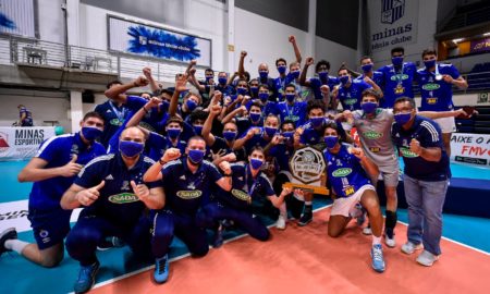Em uma quadra de vôlei, atletas e comissão técnica do Sada Cruzeiro comemoram o título do Campeonato Mineiro de Vôlei. Três atletas seguram o troféu de primeiro colocado, enquanto os demais estão com medalhas no peito. Todas usam medalhas.